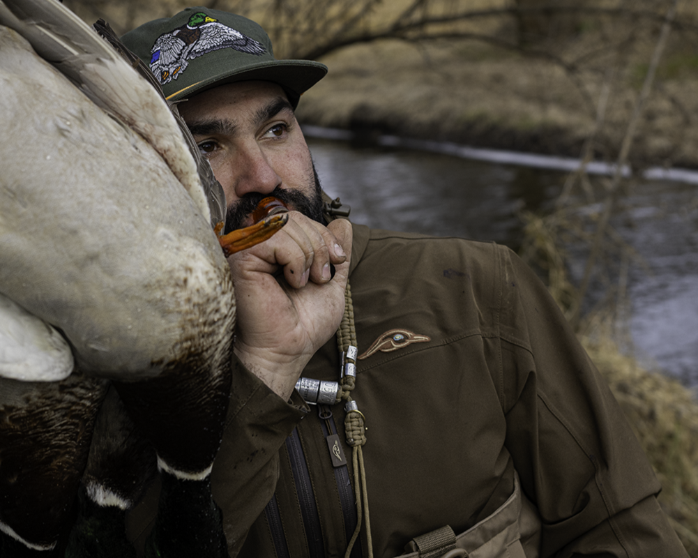 Man holding a duck near a body of water, wearing a cap and brown jacket.