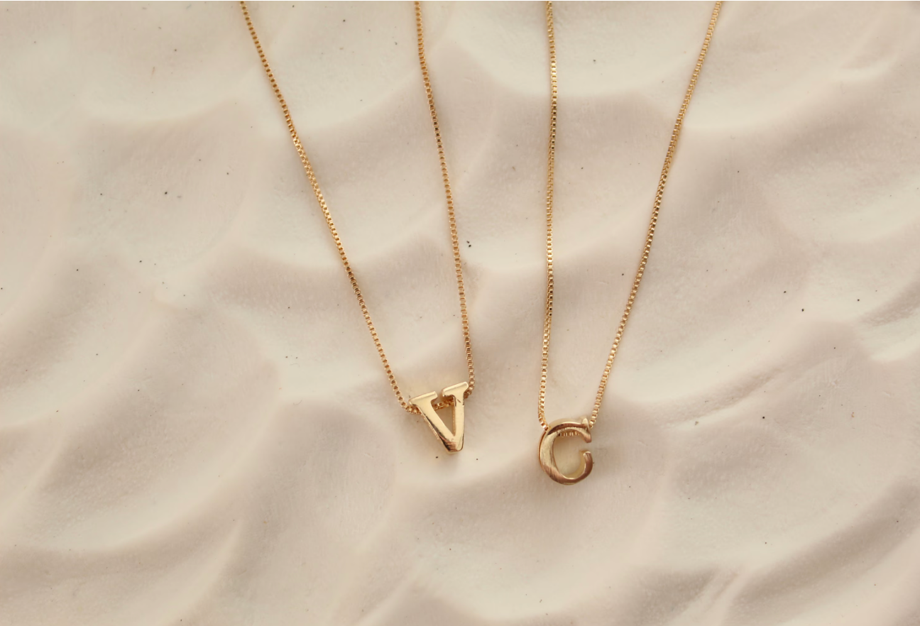 Two gold necklaces with letter pendants on a beige fabric background