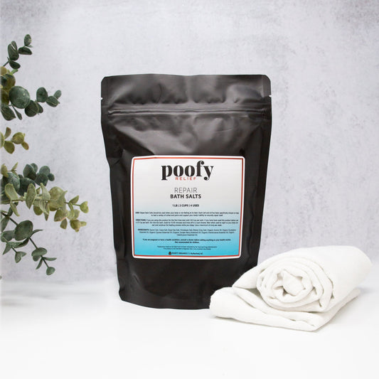 Black bag of 'poofy' cedar bath salts with a white towel and plant on a light gray background